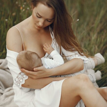 Baby wird gestillt von Frau in weißem Kleid, die im Gras sitzt stillt